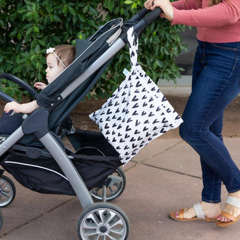 A woman pushes a stroller with a baby outdoors by greenery, with a Bumkins Wet Bag: Hearts hanging from the handle.
