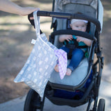 A baby sits in a stroller outdoors with a Bumkins Wet Bag: Arrow hanging from the handle, a purple cloth peeking out.
