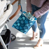 A person wearing jeans and a purple sweater places items into a Bumkins Wet Bag: Blue Tropic attached to a stroller.