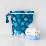 Bumkins Wet Bag: Blue Tropic sits on a white table against a white brick wall, with cloth diapers and breast pump parts in front.