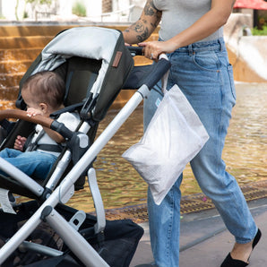 A tattooed person in jeans and a gray top pushes a stroller with a toddler by a fountain, with Bumkins Wet Bag: Wander hanging from the handle.