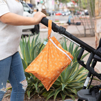 A person pushing a stroller with a Bumkins Wet Bag: Grounded attached to the handle, with plants and a street scene in the background.
