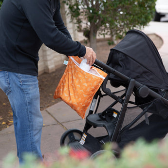 A person places a bottle into a Bumkins Wet Bag: Grounded attached to a stroller handle on a paved path with greenery.