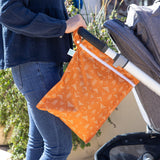 A person in jeans and a dark blue shirt pushes a stroller outdoors with a Bumkins Wet Bag: Grounded hanging from the handle.