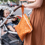 A woman in a brown dress pushes a stroller with a toddler and a Bumkins Wet Bag: Grounded hanging from the handle.