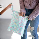 A person in a pink sweater and ripped jeans unzips a Bumkins Wet Bag: Rainbows hanging from a stroller handle.