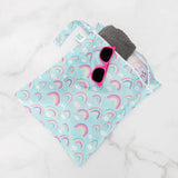 Bumkins Wet Bag: Rainbows holds a gray towel and pink sunglasses on a marble background.