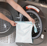 A person holds Bumkins Wet Bag: Rainbows in front of an open front-loading washing machine.