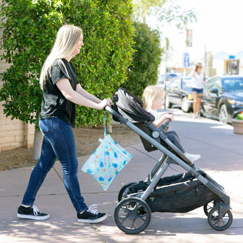 A woman pushes a child in a stroller on a sunny sidewalk, with a Bumkins Wet Bag: Ocean Life attached to the handle.