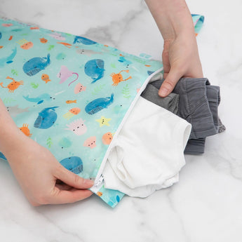 Hands placing clothes into a Bumkins Wet Bag: Ocean Life on a marble surface.