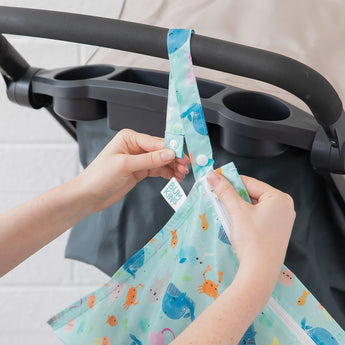 Hands attach a Bumkins Wet Bag: Ocean Life to a stroller handle using the snap button on the strap.