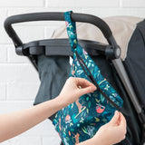 Hands unzip a Bumkins Wet Bag: Jungle, attached to a stroller handle with a white brick wall in the background.