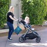 A woman pushes a stroller with a toddler inside, a Bumkins Wet Bag: Jungle hanging from the handle; trees and a building in the background.