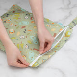 Hands zipping a Bumkins Wet Bag: Camp Gear, featuring trees and tents on waterproof fabric, on a marble surface.