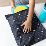 Close-up of a child placing a yellow container in a Bumkins Wet Bag: Mickey Mouse Icon Black + White next to a blue water bottle on a table.