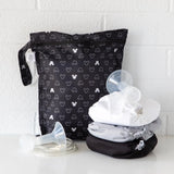 Bumkins Wet Bag: Mickey Mouse Icon Black + White sits against a white brick wall, with cloth diapers and breast pump parts in front.