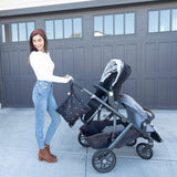 A person in a white sweater pushes a stroller by a garage, with a Bumkins Wet Bag: Mickey Mouse Icon Black + White attached to the handle.