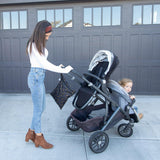 A woman pushes a child in a stroller by a garage, with a Bumkins Wet Bag: Minnie Mouse Icon Black + White hanging from the handle.