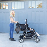 A person smiles with a child in a stroller by a blue garage, with Bumkins Wet Bag: Minnie Mouse Icon Black + White hanging from the handle.