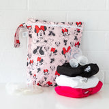 Bumkins Wet Bag: Minnie Mouse Classic sits on a white table against a white brick wall with cloth diapers and breast pump parts in front.