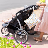 A person pushes a stroller on a sunny day with Bumkins Wet Bag: Winnie and Friends hanging from the handle, pink flowers nearby.
