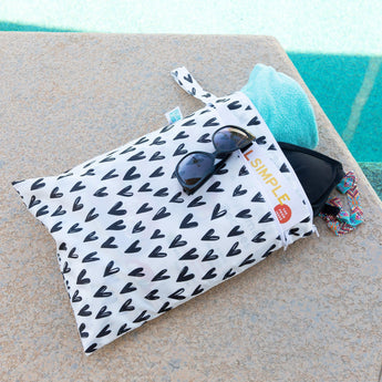 Bumkins Wet/Dry Bag: Hearts holds sunglasses, a turquoise towel, and a swimsuit by a pool.