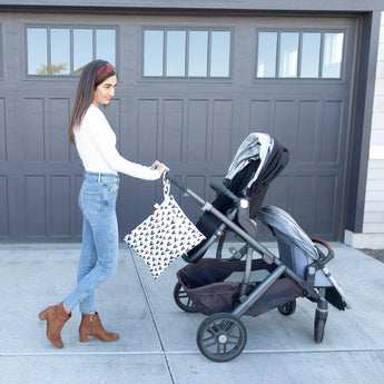 A woman wearing jeans & a white top pushes a double stroller with a Bumkins Wet/Dry Bag: Hearts attached to the handle in front of a garage.