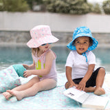 Two kids in sun hats sit by a pool on a colorful mat. One holds a book, while the other grabs a towel from a Bumkins Wet/Dry Bag: Rainbows.