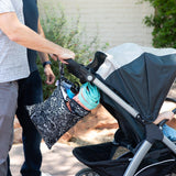 A person pushes a stroller outdoors with a Bumkins Wet/Dry Bag: Be Kind hanging from the handle; a towel and a water bottle peeking out.
