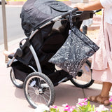 Under a sunny sky, a person in a pink skirt and sandals pushes a black stroller with a Bumkins Wet/Dry Bag: Be Kind hanging from the handle.