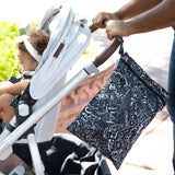 A person pushes a stroller with a child outdoors while a Bumkins Wet/Dry Bag: Be Kind hangs from the handle.