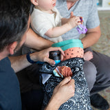 Two adults interact; one arranges items in a Bumkins Wet/Dry Bag: Be Kind, while the other holds a baby clutching a toy.