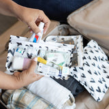 Hands fill a Bumkins Clear Travel Bag 3-Pack: Hearts with toiletries above a packed suitcase, with the other bags on top.