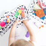 A child reaches into Bumkins Clear Travel Bag 3-Pack: Hearts filled with craft supplies on a white table.