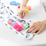 Close-up of a child's hand zipping a Bumkins Clear Travel Bag 3-Pack: Hearts filled with colorful pom-poms on a white table.