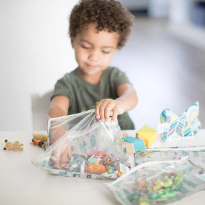 A curly-haired child opens Bumkins Clear Travel Bag 3-Pack: Cacti filled with toys in a bright room at a white table.