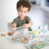 A curly-haired child opens Bumkins Clear Travel Bag 3-Pack: Cacti filled with toys in a bright room at a white table.