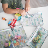 A child sits with Bumkins Clear Travel Bag 3-Pack: Cacti filled with toys on a white table.