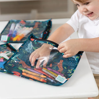 A child smiles while organizing markers in Bumkins Clear Travel Bag 3-Pack: Jungle on a white table.