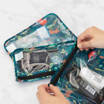 Hands unzip Bumkins Clear Travel Bag 3-Pack: Jungle filled with toiletries and makeup on a marble surface.