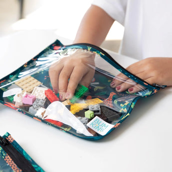 A child's hand reaches for colorful toy brick pieces from a Bumkins Clear Travel Bag 3-Pack: Jungle on a white table.
