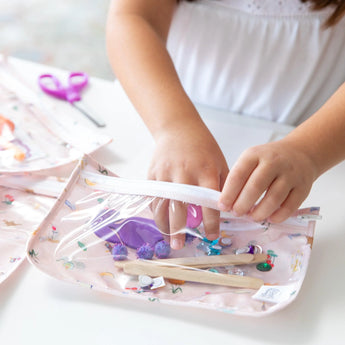 A child expertly packs craft supplies into Bumkins Clear Travel Bag 3 Pack: Princess Magic at a white table.