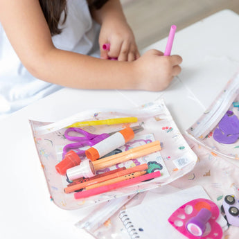 A child draws with a pink marker beside Bumkins Clear Travel Bag 3 Pack: Princess Magic filled with art supplies.