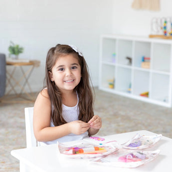 A young girl with long hair smiles at a white table, with art supplies in Bumkins Clear Travel Bag 3 Pack: Princess Magic nearby.