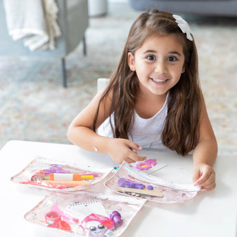 A young girl smiles at a white table with art supplies in Bumkins Clear Travel Bag 3 Pack: Princess Magic.