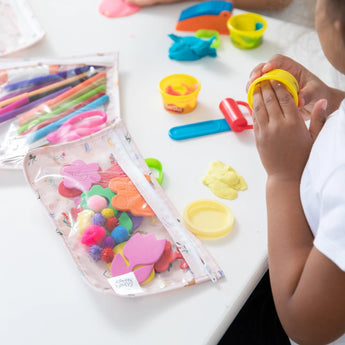 A child enjoys modeling clay on a white table, with craft supplies stored in Bumkins Clear Travel Bag 3 Pack: Princess Magic.