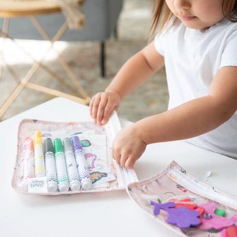 A child organizes markers and stickers in a Bumkins Clear Travel Bag 3 Pack: Princess Magic on a white table.