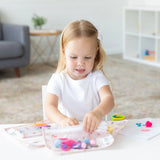 A young girl crafts happily at a white table, surrounded by Bumkins Clear Travel Bag 3 Pack: Princess Magic filled with art supplies.