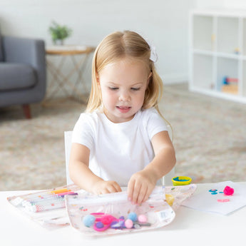 A young girl crafts happily at a white table, surrounded by Bumkins Clear Travel Bag 3 Pack: Princess Magic filled with art supplies.