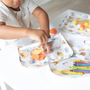 A child crafts joyfully at a white table with colorful supplies in Bumkins Clear Travel Bag 3-Pack: Winnie and Friends.